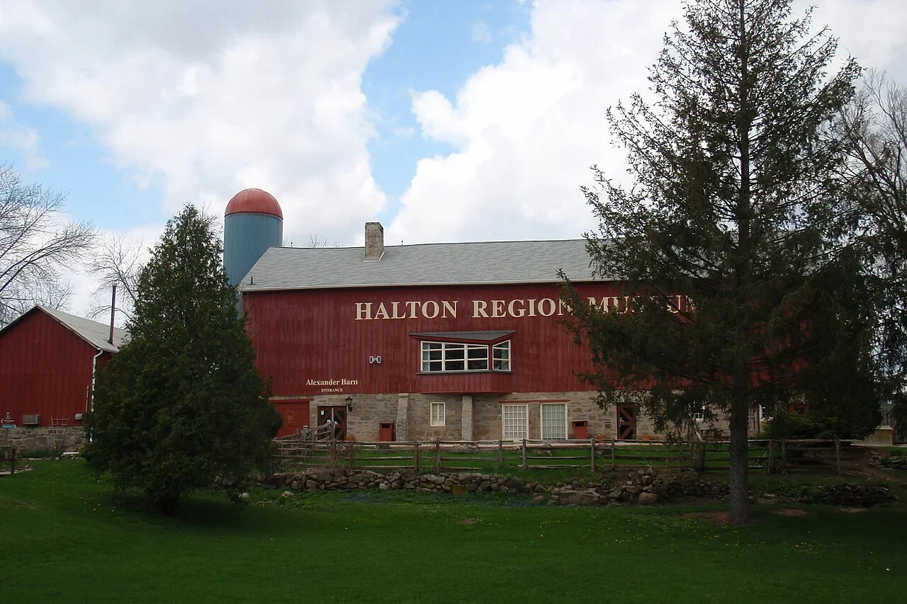 Red barn on green lawn with large trees - sign on barn says "Halton Region Museum"